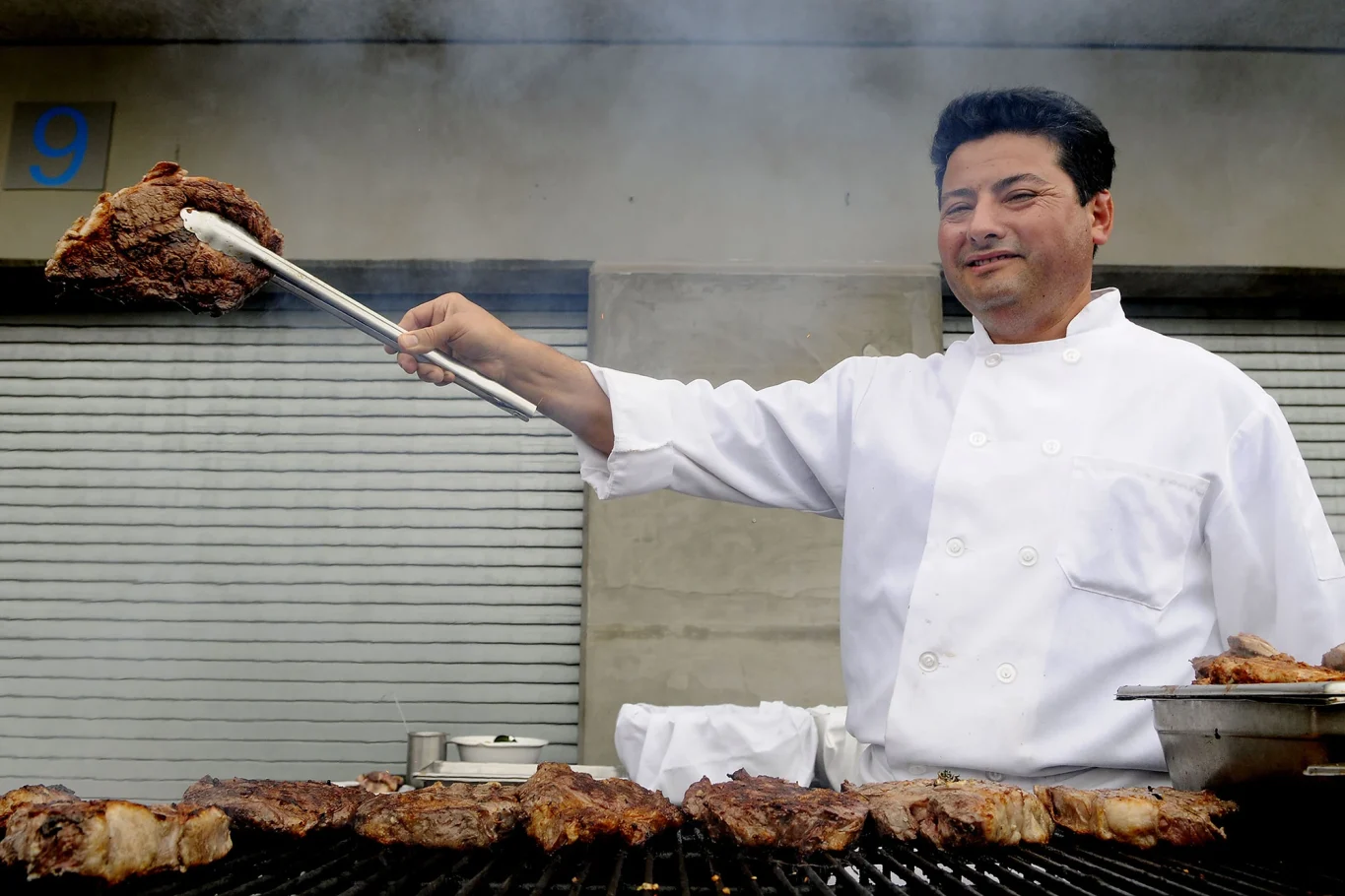 Grillmeister zeigt Fleischstücke mit einer Zange über dem Grill.