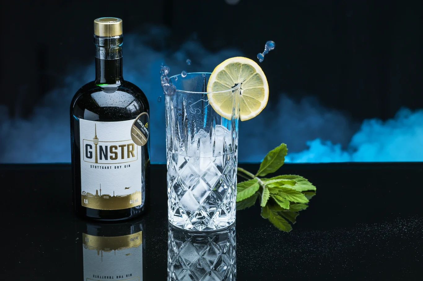 Ein Glas mit Eis, Zitrone und Gin, daneben eine Flasche Gin auf dunklem Hintergrund.