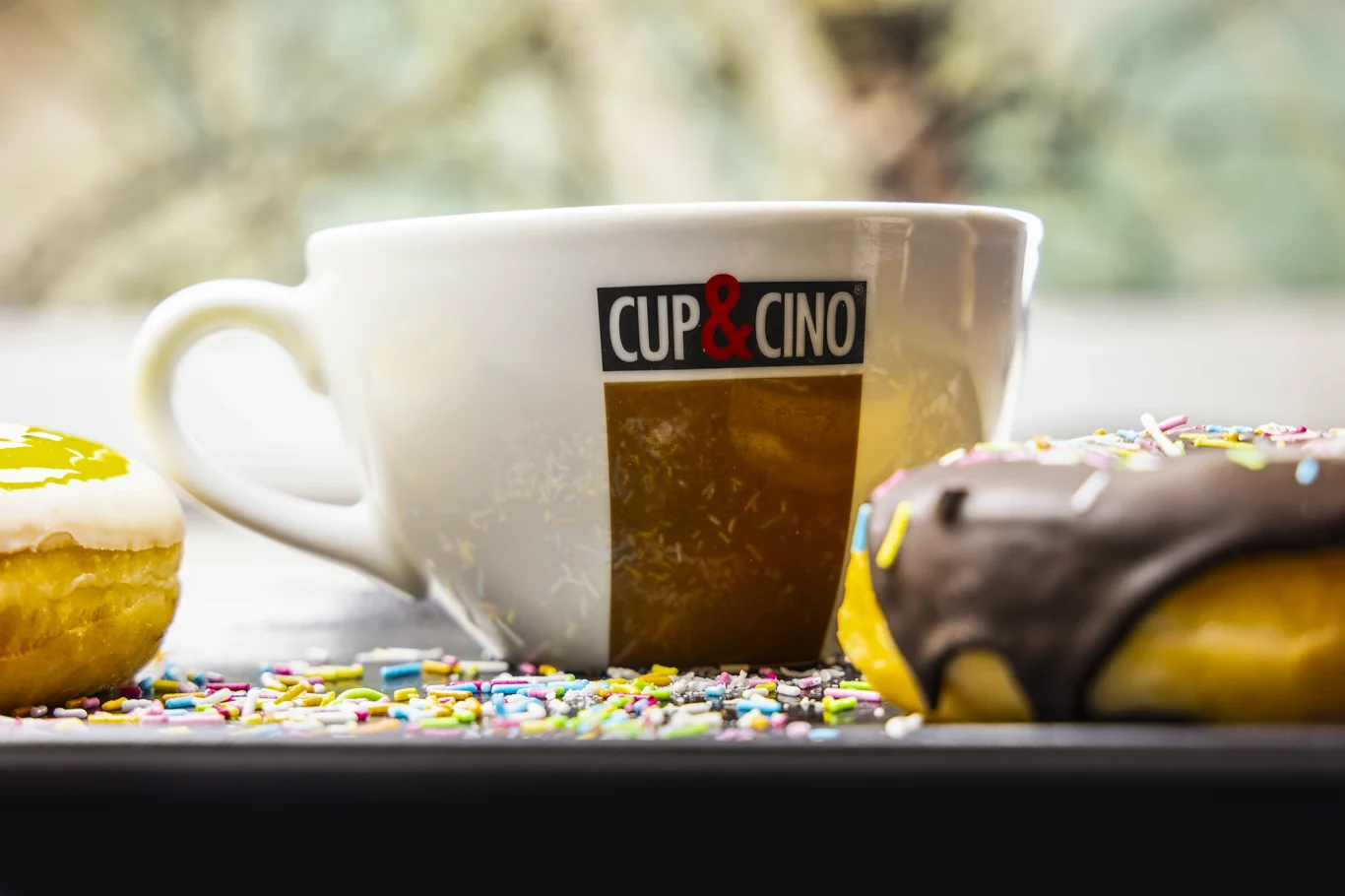 Eine Tasse mit "Cup & Co." und zwei Donuts, einer mit Schokolade und Streuseln.